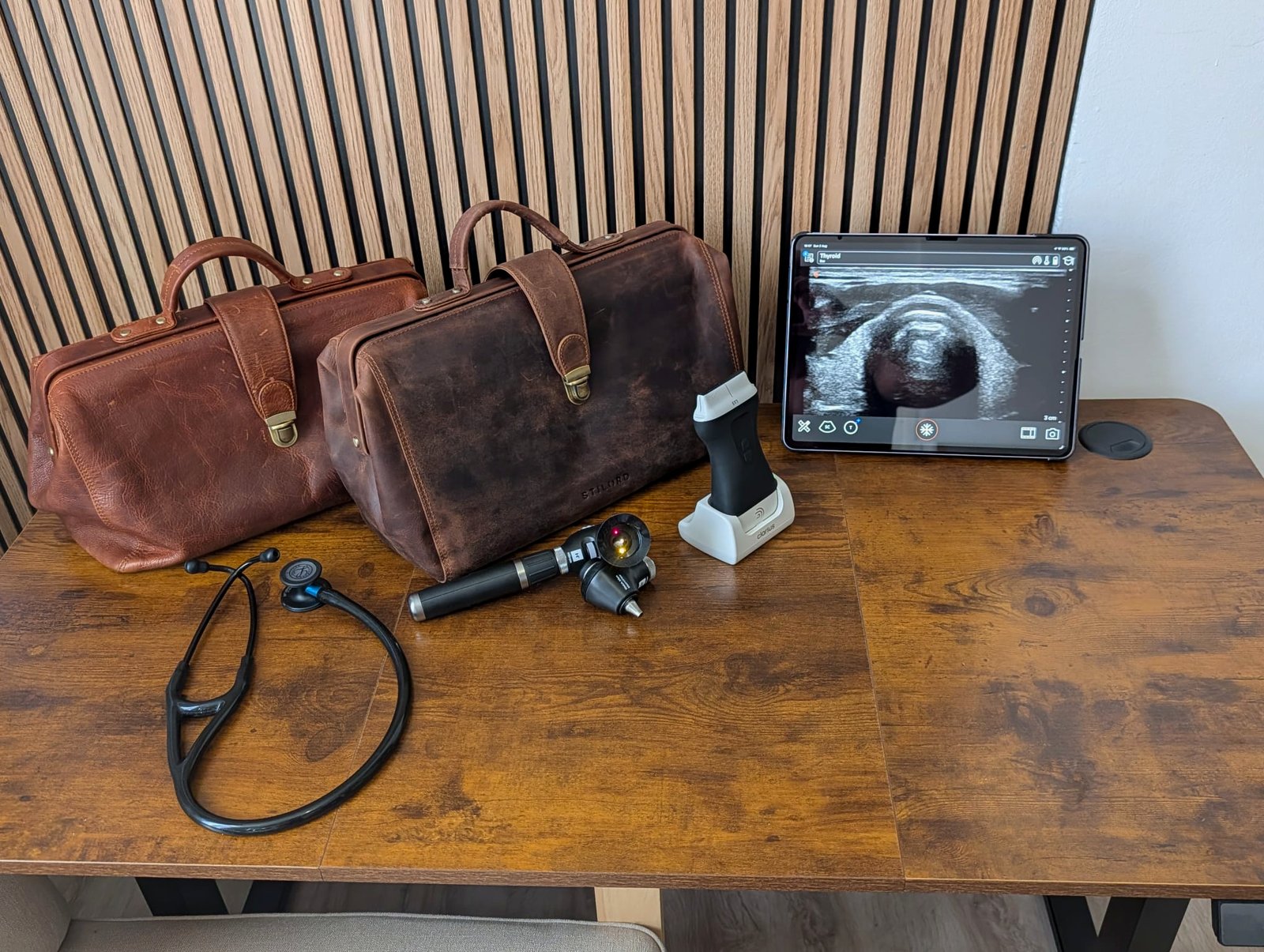 Two leather doctor bags, a stethoscope, an otoscope and ophthalmoscope set, a wireless ultrasound probe and a tablet showing a thyroid scan, on a wooden table