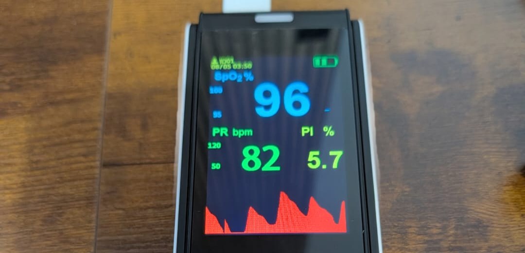 Handheld patient monitor on a table displaying oxygen saturation, pulse rate, perfusion index and waveform